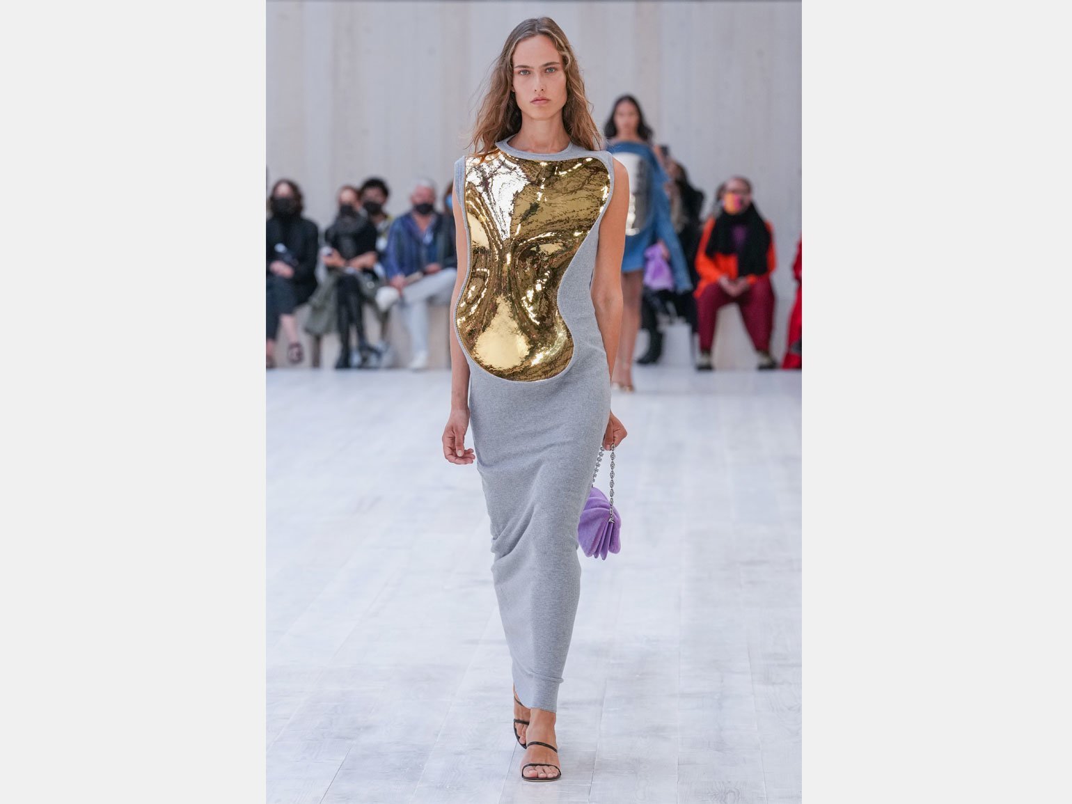 модель идёт по подиуму на показе Loewe Spring/Summer 2022 в сером платье с золотой скульптурной вставкой спереди и с бархатной сиреневой сумкой на цепочке