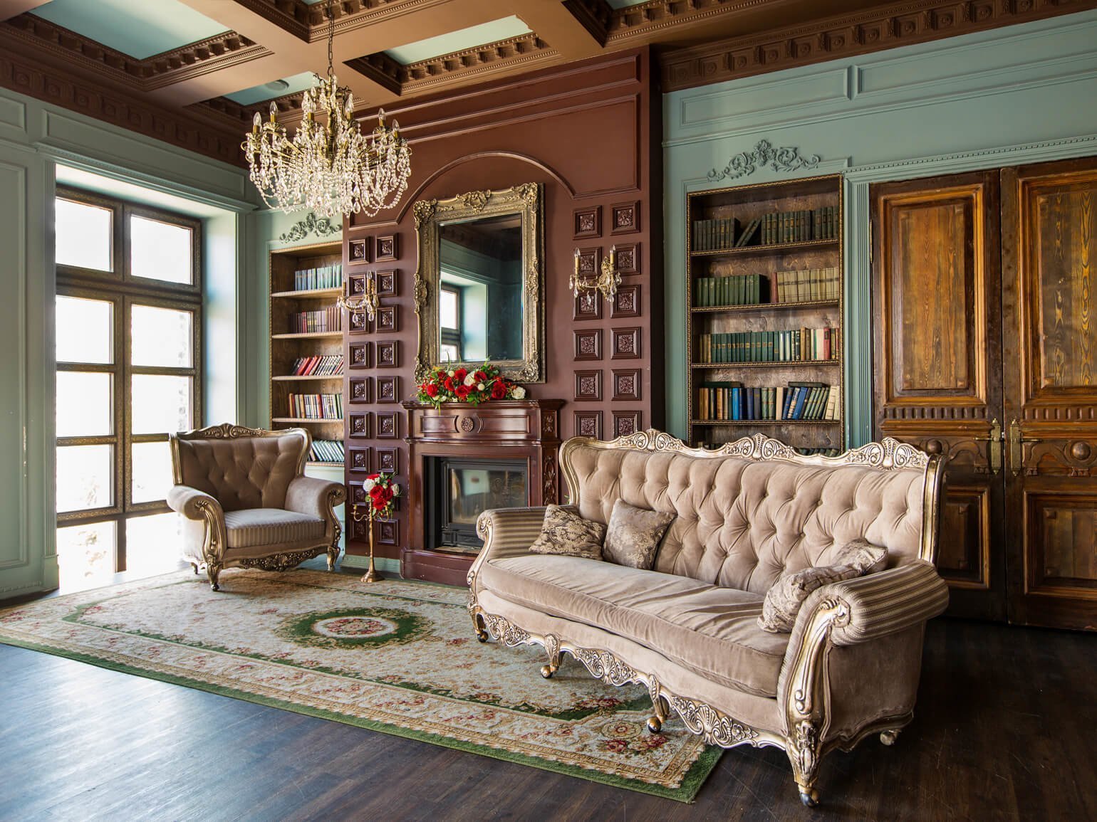 В комнате с камином бархатный диван и бархатное кресло цвета нюд с отделкой деревом золотого цвета, хрустальная люстра, персидский ковер, зеркало в золотой раме и стеллаж с книгами — дизайн интерьера в викторианском стиле