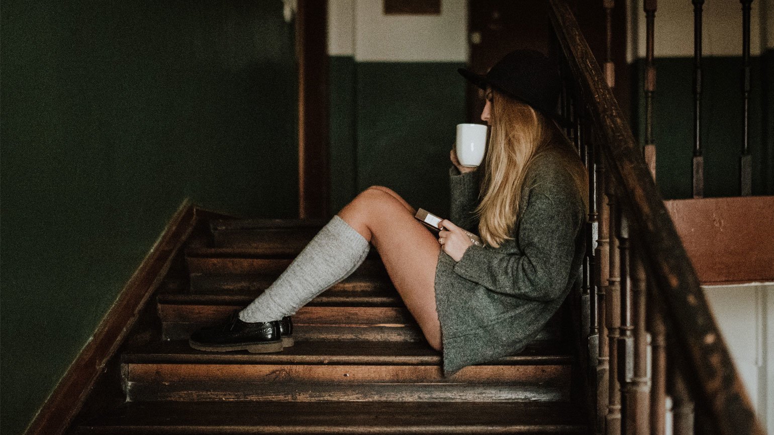 Девушка с длинными волосами в пальто и шляпе с кружкой кофе и книгой сидит на деревянной лестнице