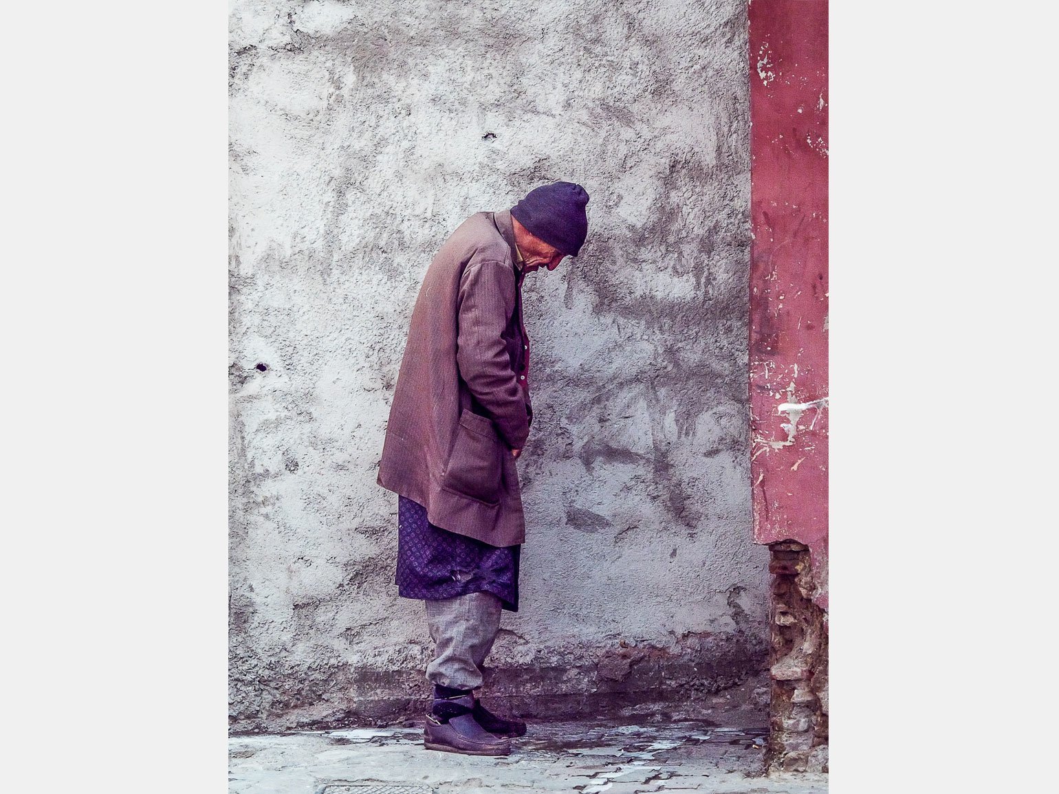 Старый мужчина в грязной одежде и синей шапке согнувшись стоит у стены