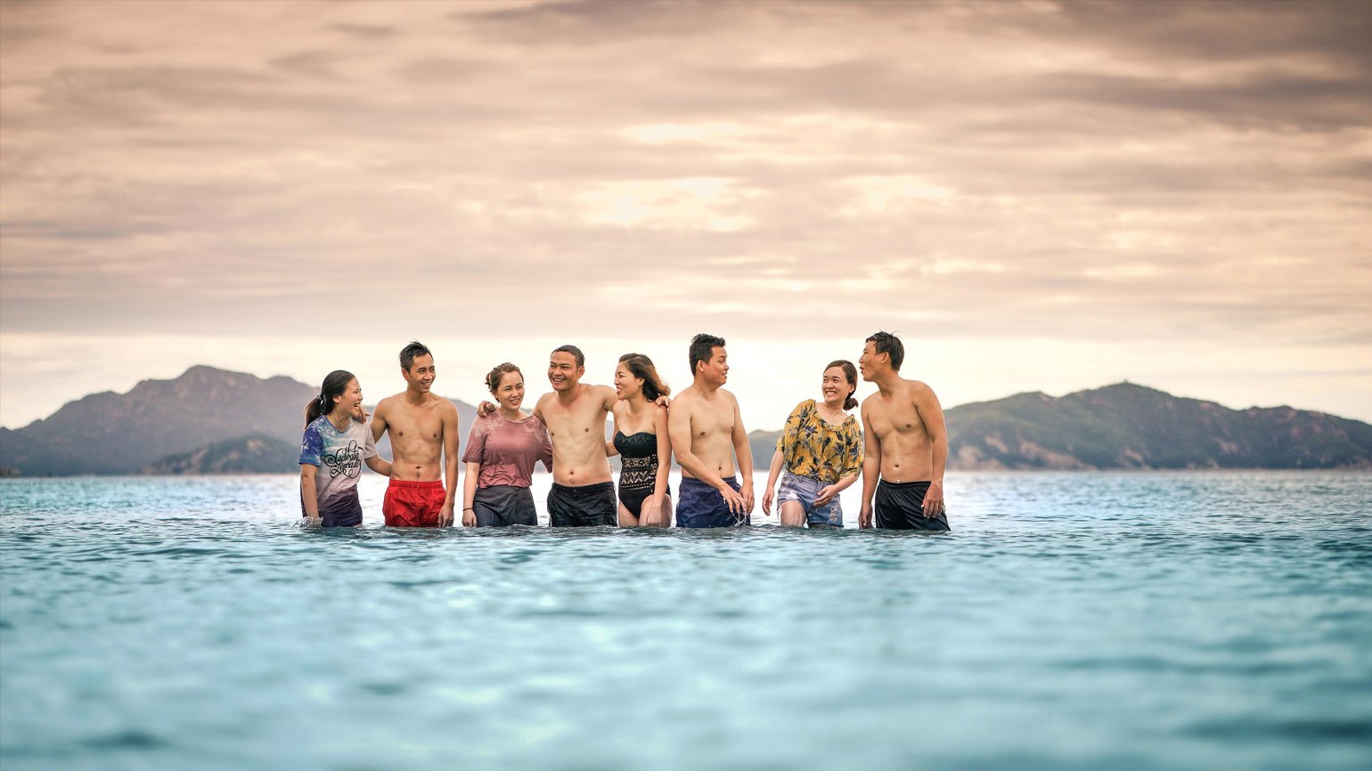 Восемь человек стоят по бёдра в морской воде на фоне гор и заката