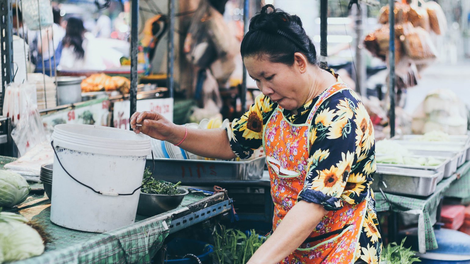 Женщина в фартуке на рынке в Таиланде перебирает зелень в вёдрах